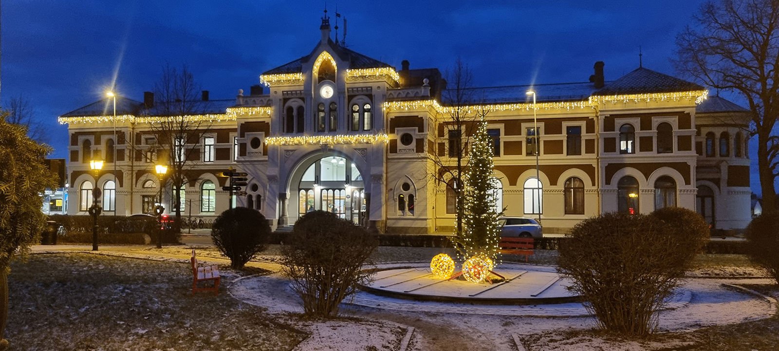 Kveldsbilde av stasjonen med julebelysning. I forgrunnen stasjonsparken med et juletre.