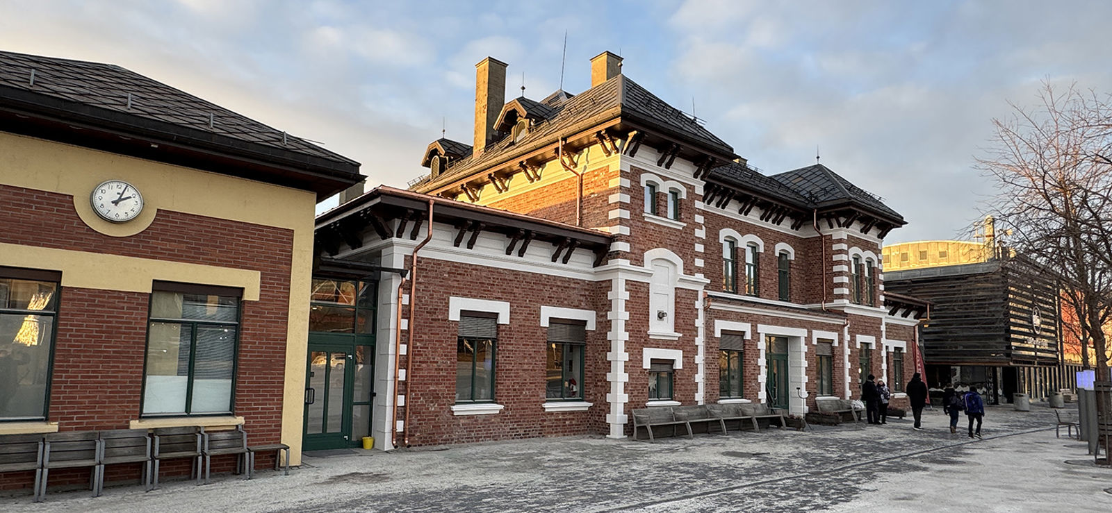 Stasjonsbygningen med noen personer og den moderne skysstasjonen i bakgrunnen.