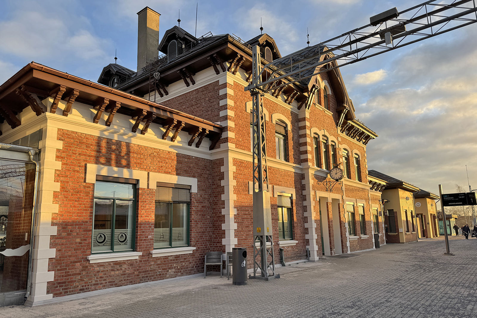 Stasjonsbygningen med plattformen i forgrunnen, åk for kontaktledningen og reisende i bakgrunnen.