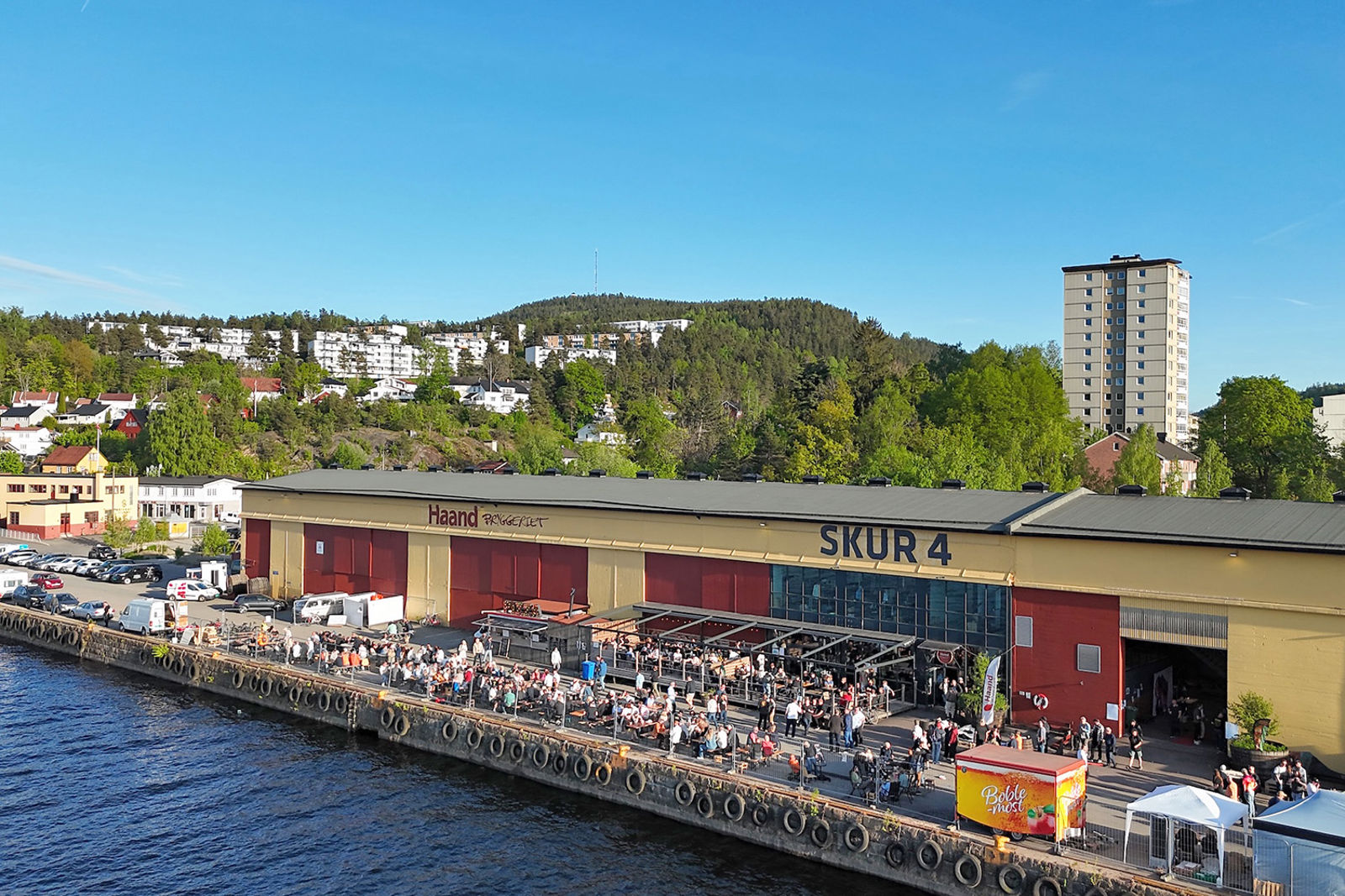 Ombygd lagerbygning langs kaia, mennesker i sola på uteservering. Annen bebyggelse i bakgrunnen.