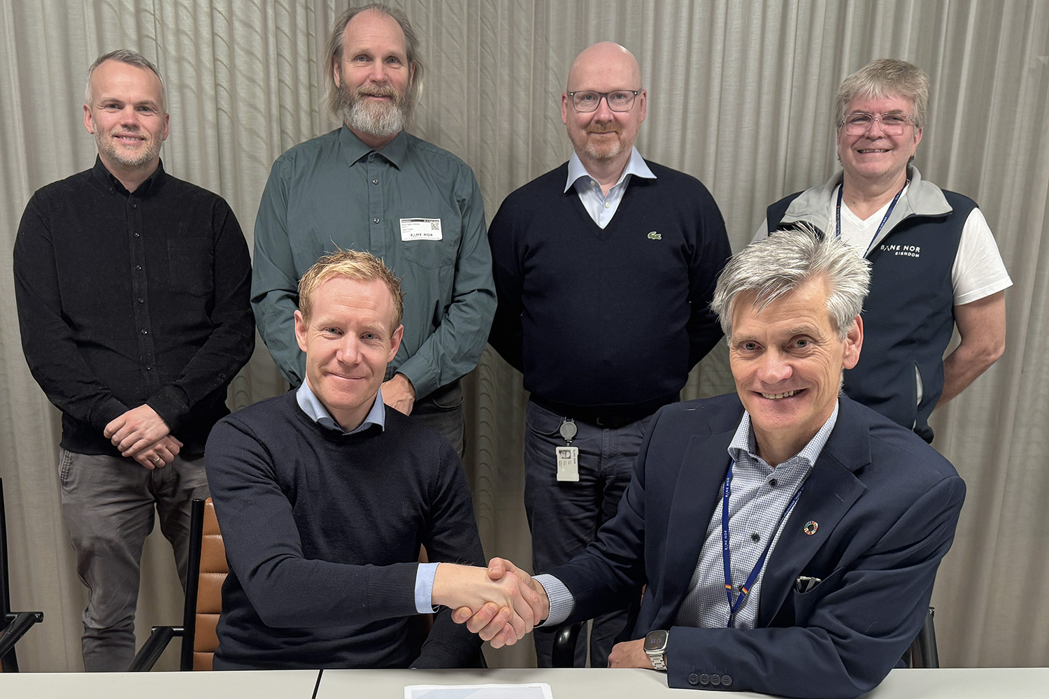 De to personene foran utveksler håndtrykk, øvrige står i en halvsirkel bak.