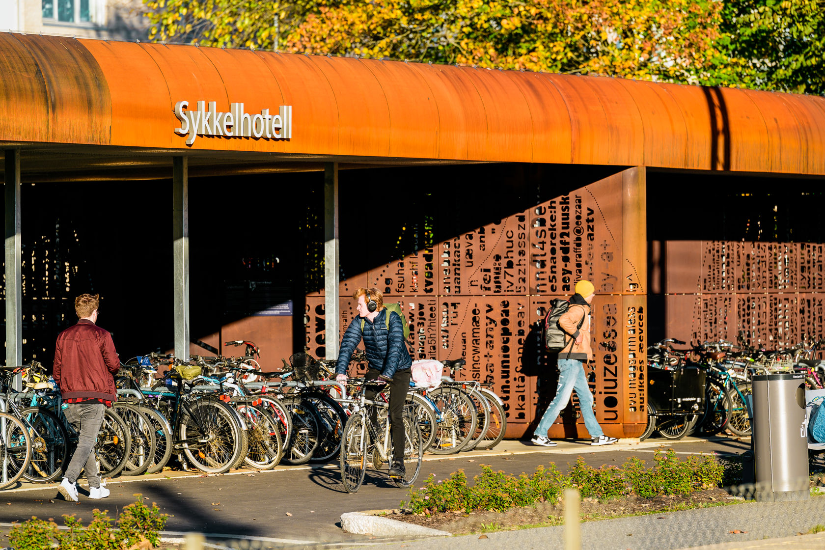 Mennesker ved sykkelhotellet i Ås. Sola skinner på sykkelhotellet som blir oransje i sollyset.