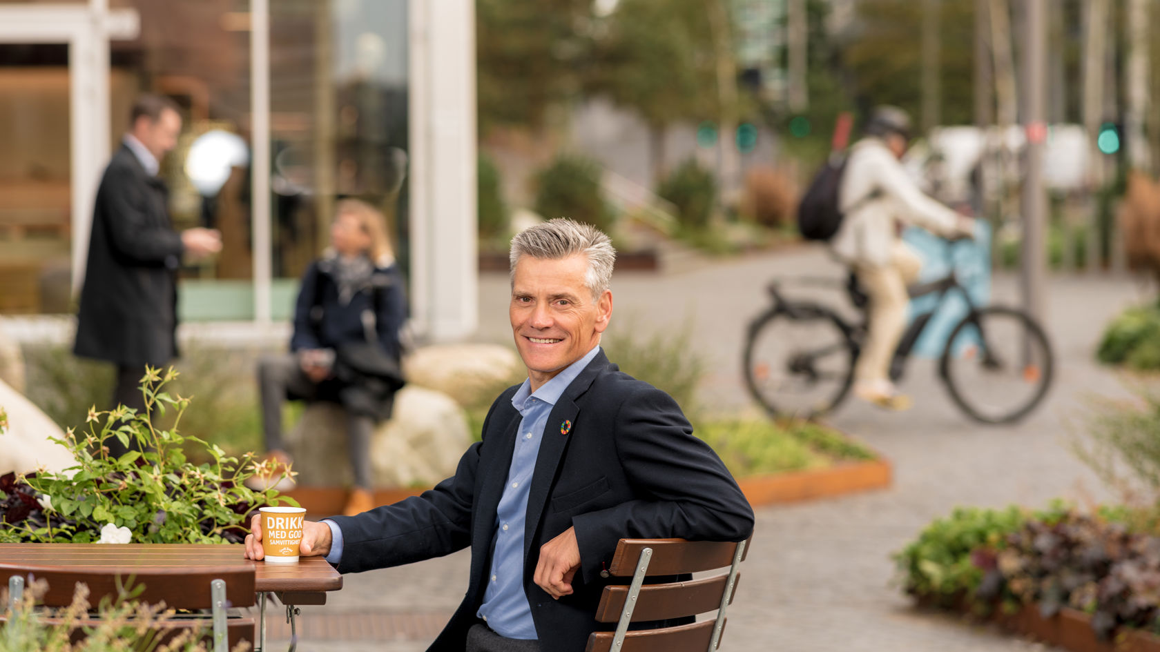 Mann i dressjakke sitter ved et cafe-bord ute og ser i kamera