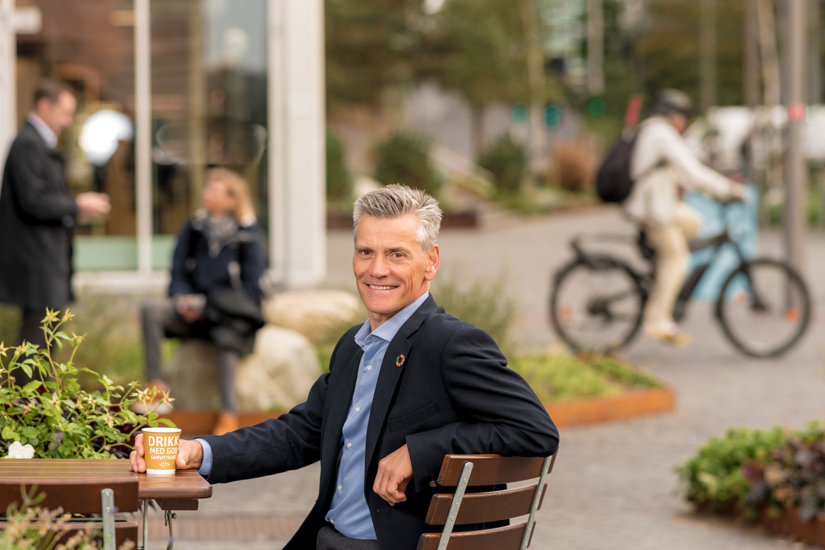 Mann i dressjakke sitter ved et cafe-bord ute og ser i kamera