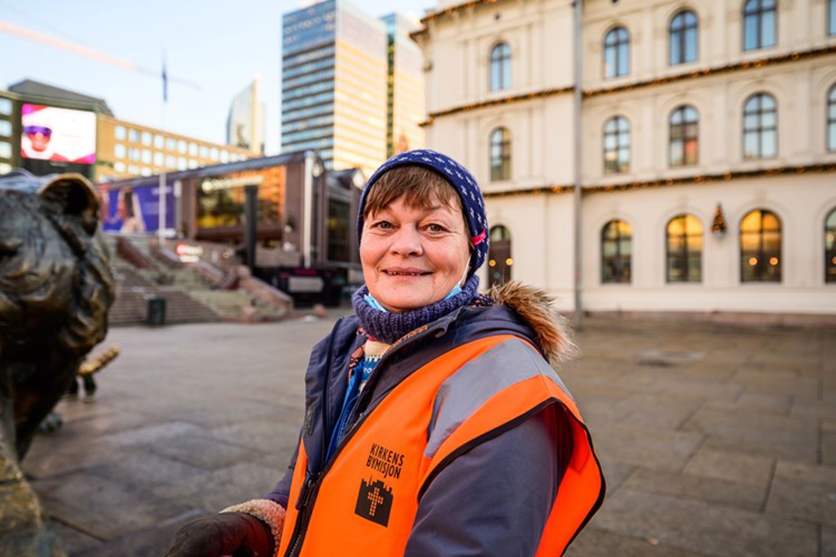 Dame i oransje vest utenfor Østbanehallen. Hun smiler, har på seg lue og boblejakke.