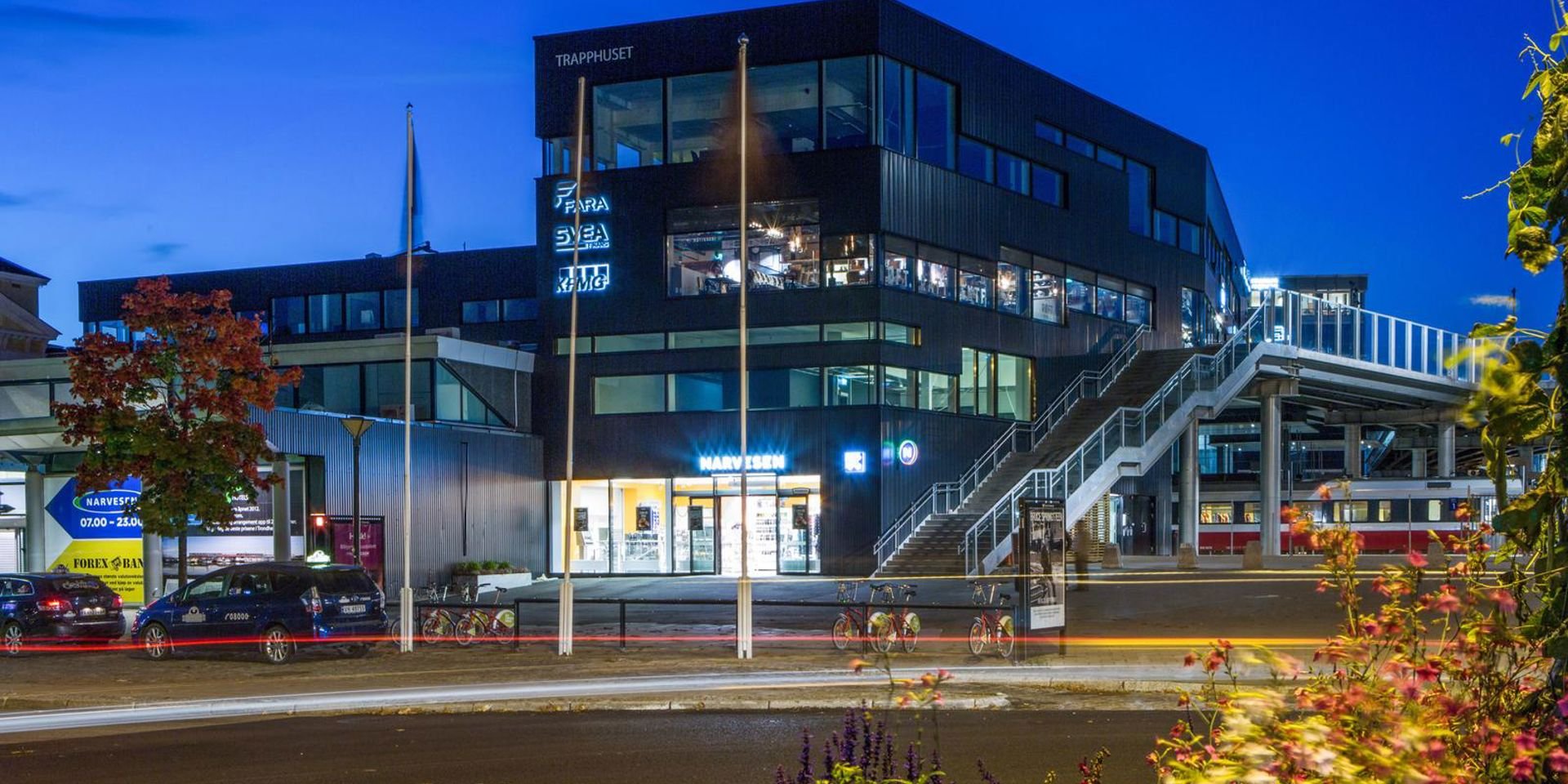 Trær i forgrunn, mørket har senket seg over Trondheim sentralstasjon som lyser opp.